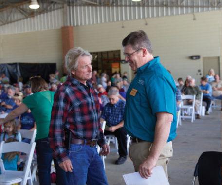 Two men talking with people sitting in rows of seats behind them.