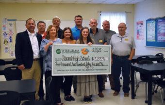 A group of people stands in front of a classroom, holding a large check, smiling and celebrating their achievement.
